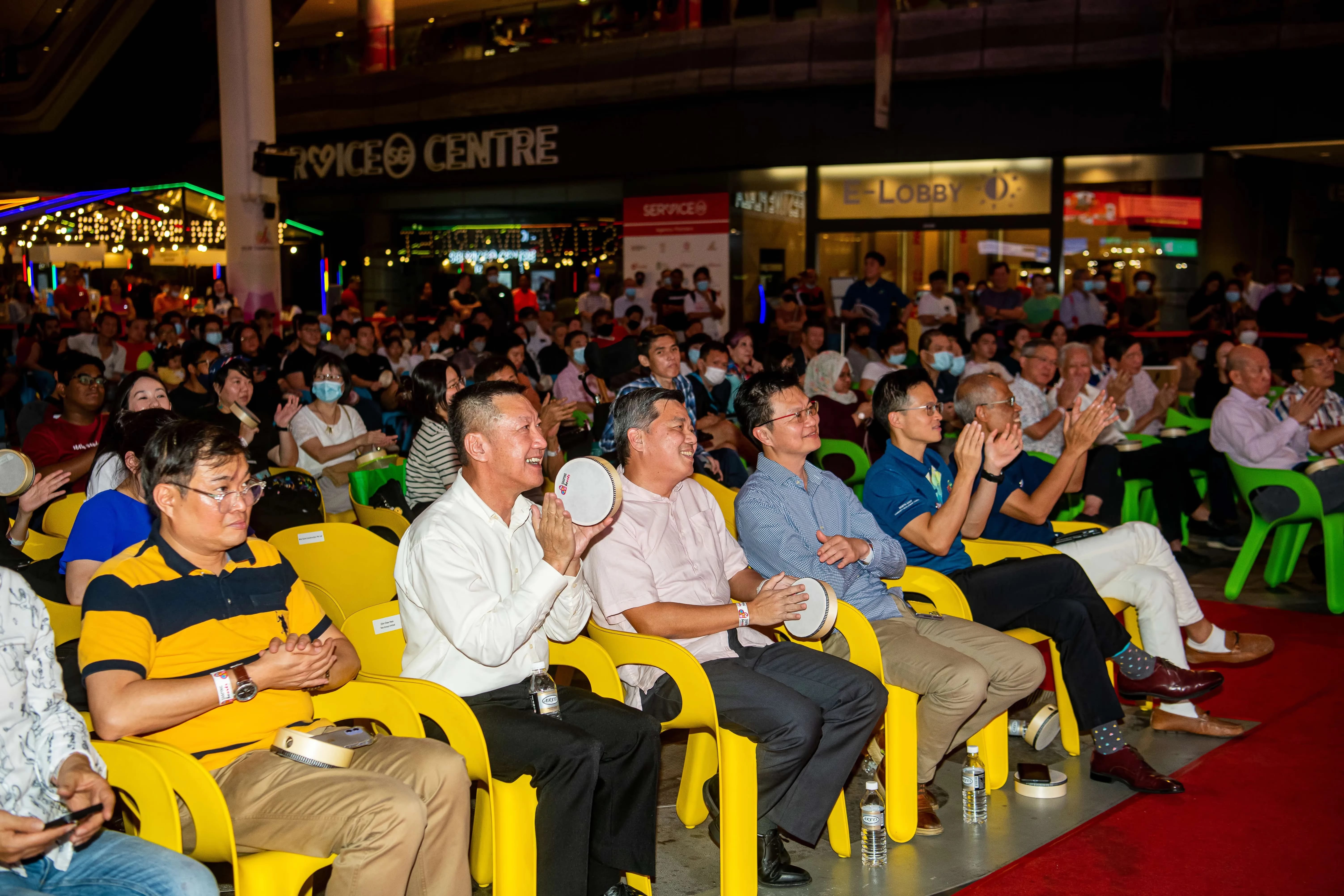 A large group of people is seated in bright yellow chairs, some holding tambourines and clapping in front of a service center.